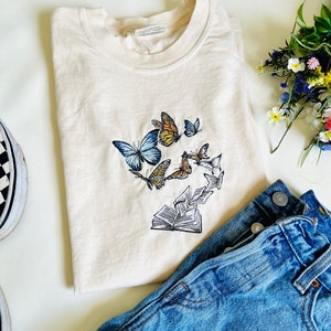 May include: A cream colored t-shirt with an embroidered design of butterflies and an open book. The butterflies are blue, yellow, and orange. The book is white and has pages that are curled up.