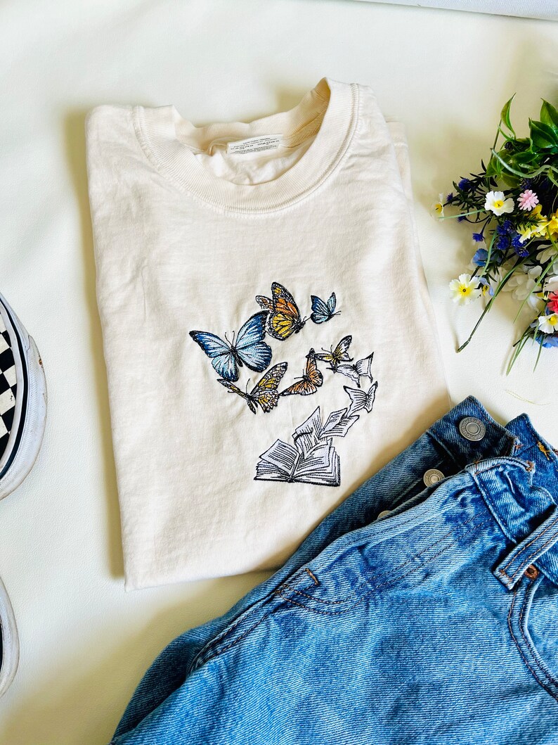 May include: A cream colored t-shirt with an embroidered design of butterflies and an open book. The butterflies are blue, yellow, and orange. The book is white and has pages that are curled up.