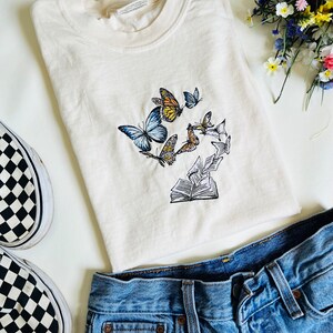 May include: A cream colored t-shirt with an embroidered design of butterflies flying around an open book. The t-shirt is paired with blue jeans and black and white checkered Vans.