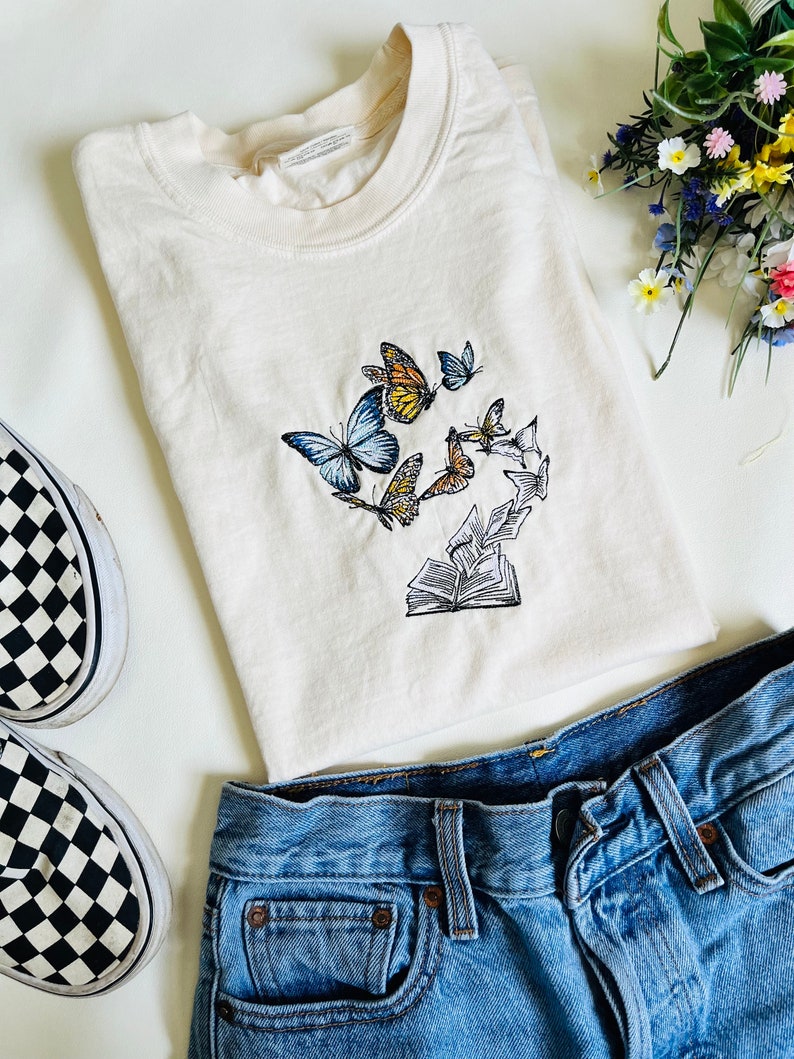 May include: A cream colored t-shirt with an embroidered design of butterflies flying around an open book. The t-shirt is paired with blue jeans and black and white checkered Vans.