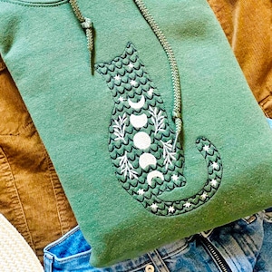 May include: A green hooded sweatshirt with a black cat design. The cat is outlined in black and has a white moon and star pattern.