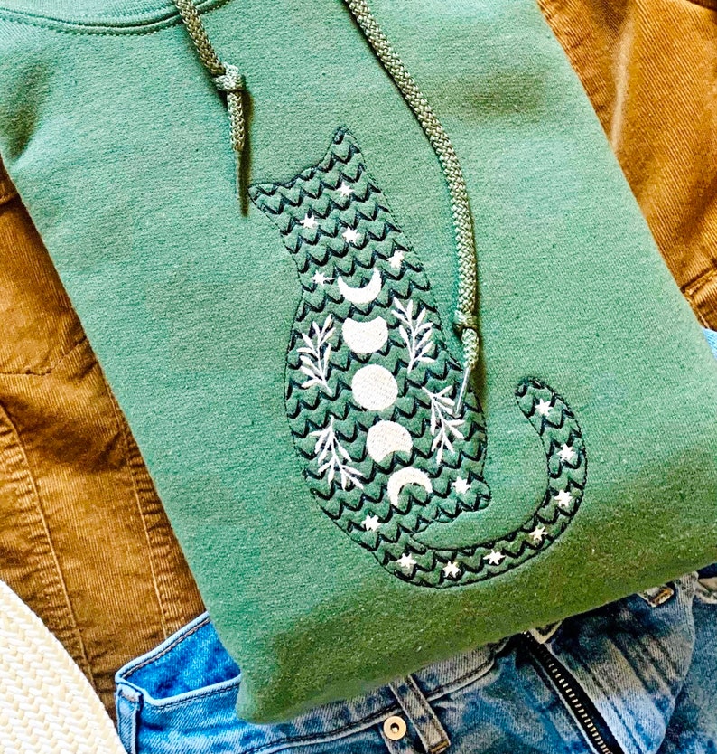 May include: A green hooded sweatshirt with a black cat design. The cat is outlined in black and has a white moon and star pattern.