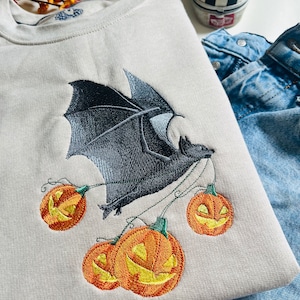 Puede incluir: Una sudadera gris con un diseño bordado de un murciélago que lleva una cadena de calabazas. El murciélago es negro y las calabazas son naranjas con caras negras.