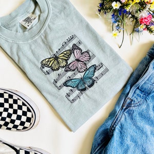 May include: A light blue t-shirt with a design of three butterflies on a musical staff. The butterflies are yellow, pink, and blue.