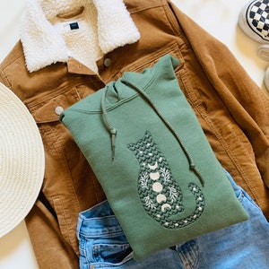 May include: A green hooded sweatshirt with a white embroidered cat design featuring a crescent moon and stars. The sweatshirt is laid on top of a brown corduroy jacket and blue jeans.