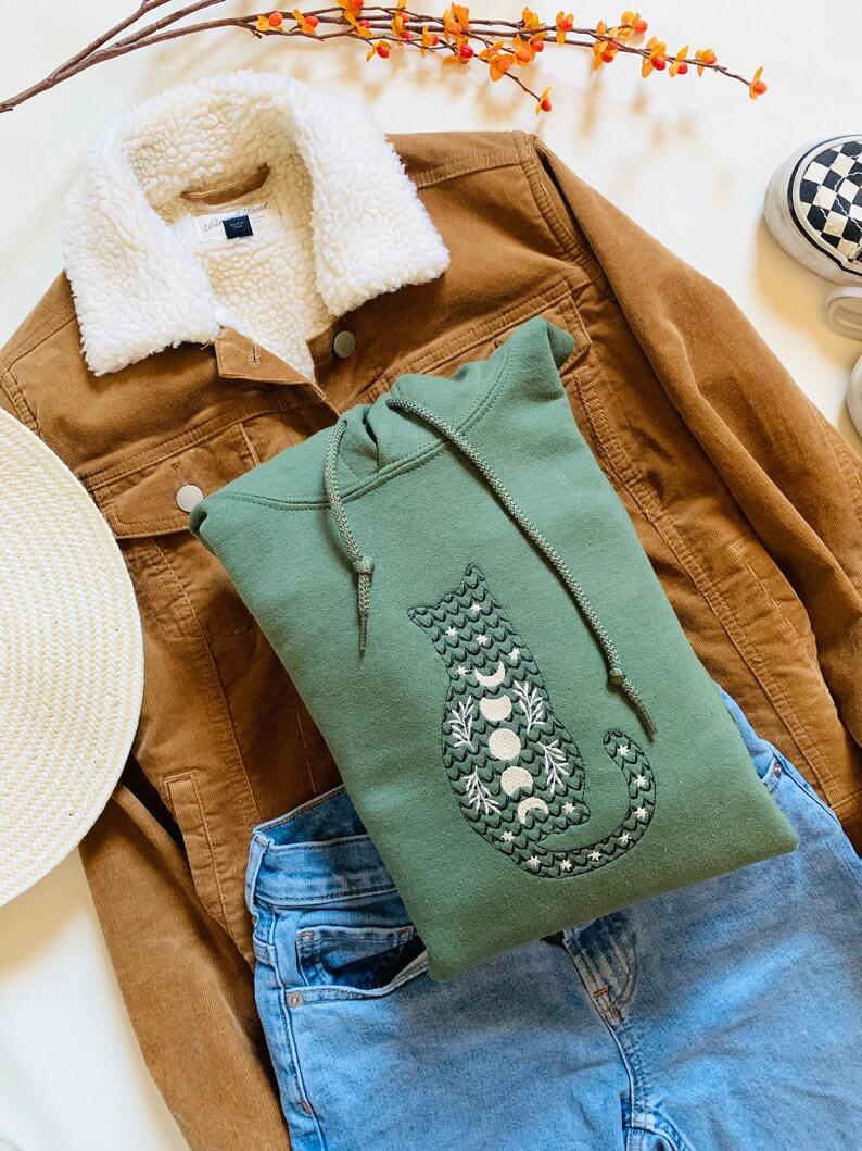 May include: A green hooded sweatshirt with a white embroidered cat design featuring a crescent moon and stars. The sweatshirt is laid on top of a brown corduroy jacket and blue jeans.