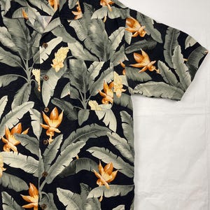 Puede incluir: Camisa de manga corta con botones y estampado floral tropical. La camisa presenta un fondo negro con hojas grises grandes y flores naranjas y amarillas. Tiene un bolsillo en el pecho y botones de madera.