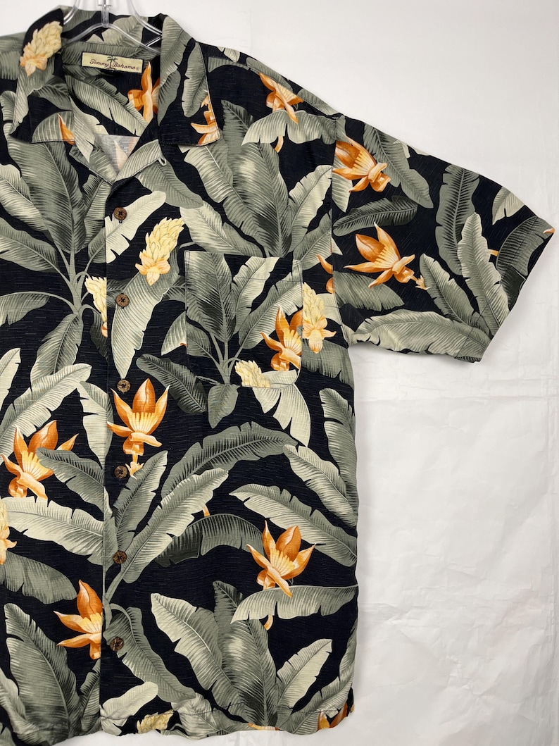 Puede incluir: Camisa de manga corta con botones y estampado floral tropical. La camisa presenta un fondo negro con hojas grises grandes y flores naranjas y amarillas. Tiene un bolsillo en el pecho y botones de madera.