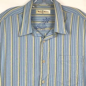 Puede incluir: Camisa abotonada con rayas verticales en azul claro, amarillo y blanco. La camisa tiene un bolsillo en el pecho y un cuello. La etiqueta muestra la marca "Tommy Bahama".