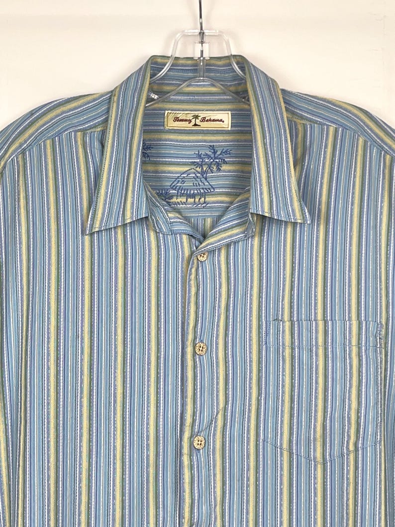 Puede incluir: Camisa abotonada con rayas verticales en azul claro, amarillo y blanco. La camisa tiene un bolsillo en el pecho y un cuello. La etiqueta muestra la marca "Tommy Bahama".
