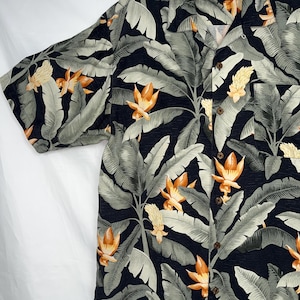 Puede incluir: Camisa de manga corta con estampado floral tropical. La camisa tiene un fondo azul marino oscuro con grandes hojas de pl&aacute;tano verde claro y flores naranjas y amarillas. Tiene un cuello cl&aacute;sico y botones de madera.