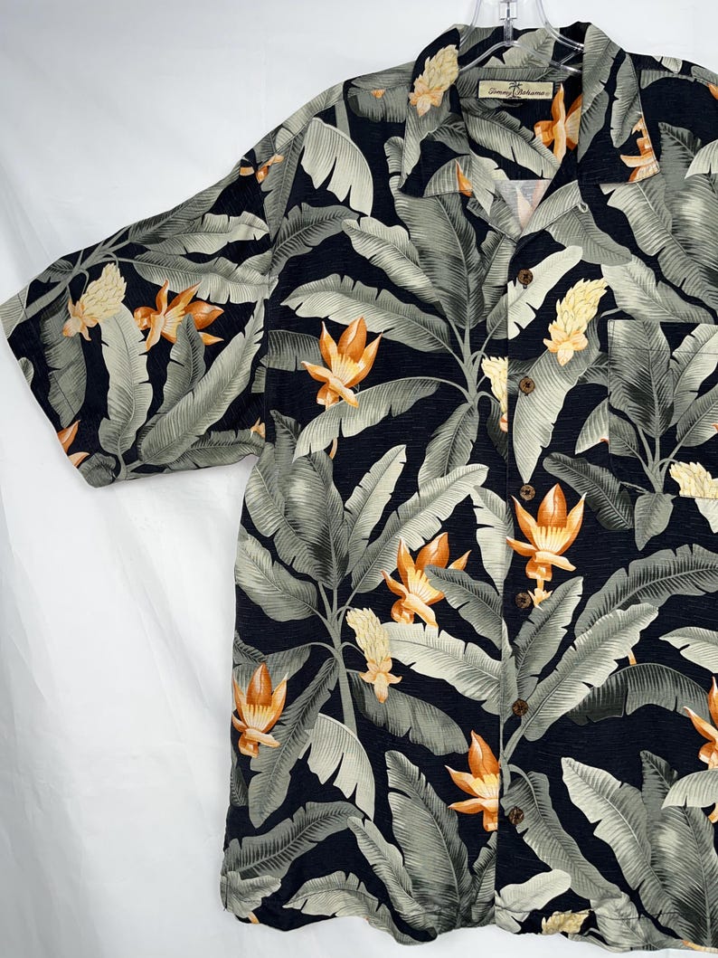 Puede incluir: Camisa de manga corta con estampado floral tropical. La camisa tiene un fondo azul marino oscuro con grandes hojas de pl&aacute;tano verde claro y flores naranjas y amarillas. Tiene un cuello cl&aacute;sico y botones de madera.