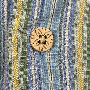 Puede incluir: Primer plano de una camisa abotonada. La camisa presenta rayas verticales en azul, blanco, verde y amarillo. Un bot&oacute;n redondo de madera con un dise&ntilde;o floral tallado est&aacute; abrochado en la tela azul.