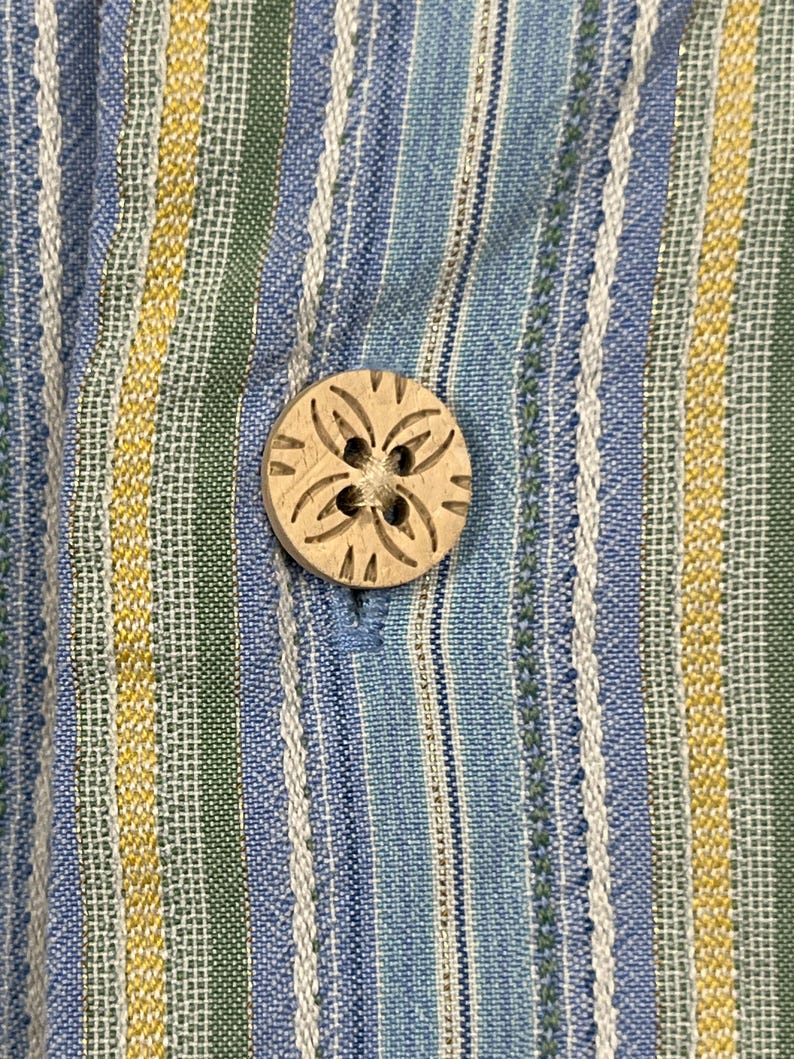 Puede incluir: Primer plano de una camisa abotonada. La camisa presenta rayas verticales en azul, blanco, verde y amarillo. Un bot&oacute;n redondo de madera con un dise&ntilde;o floral tallado est&aacute; abrochado en la tela azul.