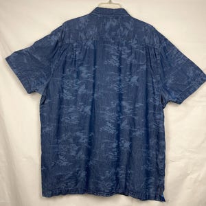 Może przedstawiać: Koszula z kr&oacute;tkim rękawem z dżinsu w kolorze średniego błękitu, zapinana na guziki. Koszula ma subtelny, całopowierzchniowy wz&oacute;r z ciemniejszego tropikalnego listowia. Wisi na białym wieszaku.