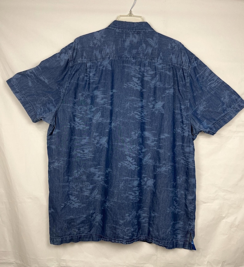 Może przedstawiać: Koszula z kr&oacute;tkim rękawem z dżinsu w kolorze średniego błękitu, zapinana na guziki. Koszula ma subtelny, całopowierzchniowy wz&oacute;r z ciemniejszego tropikalnego listowia. Wisi na białym wieszaku.