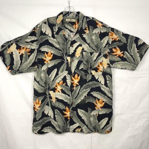 Puede incluir: Camisa de manga corta con estampado de hojas y flores tropicales. La camisa presenta un fondo oscuro con hojas grises grandes y flores naranjas. La camisa est&aacute; hecha de tela ligera y tiene un cuello cl&aacute;sico.