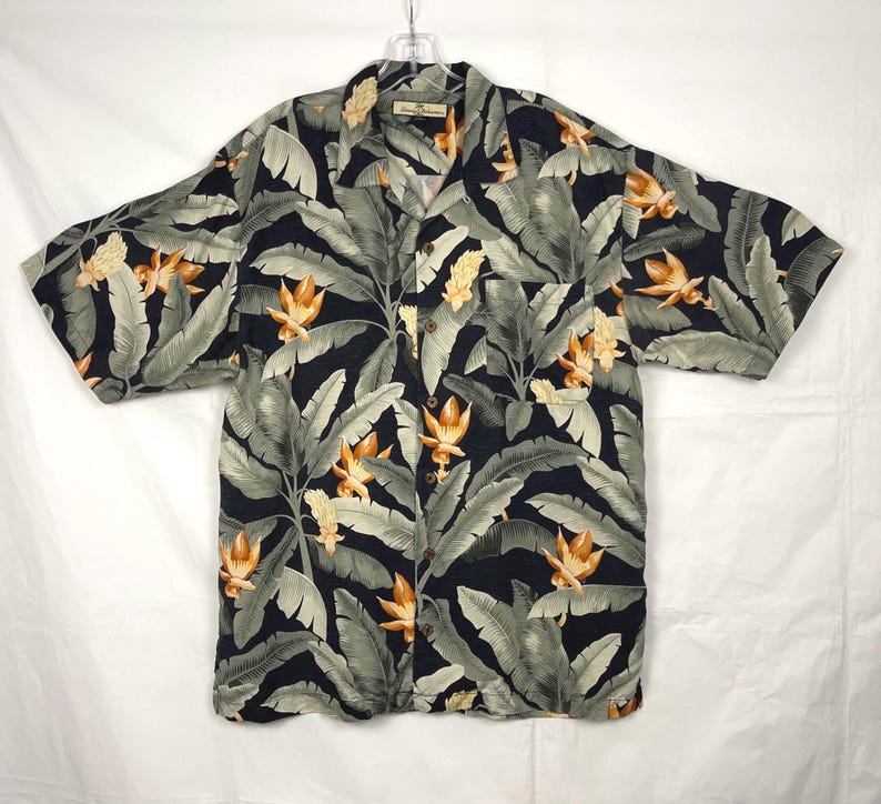 Puede incluir: Camisa de manga corta con estampado de hojas y flores tropicales. La camisa presenta un fondo oscuro con hojas grises grandes y flores naranjas. La camisa est&aacute; hecha de tela ligera y tiene un cuello cl&aacute;sico.
