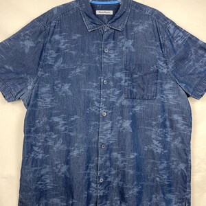 Może przedstawiać: Koszula dżinsowa z kr&oacute;tkim rękawem w średnim niebieskim odcieniu, z subtelnym kwiatowym wzorem w jaśniejszym odcieniu niebieskiego. Posiada jedną kieszeń na piersi i klasyczny kołnierzyk. Koszula jest zapięta i wisi na białym wieszaku.