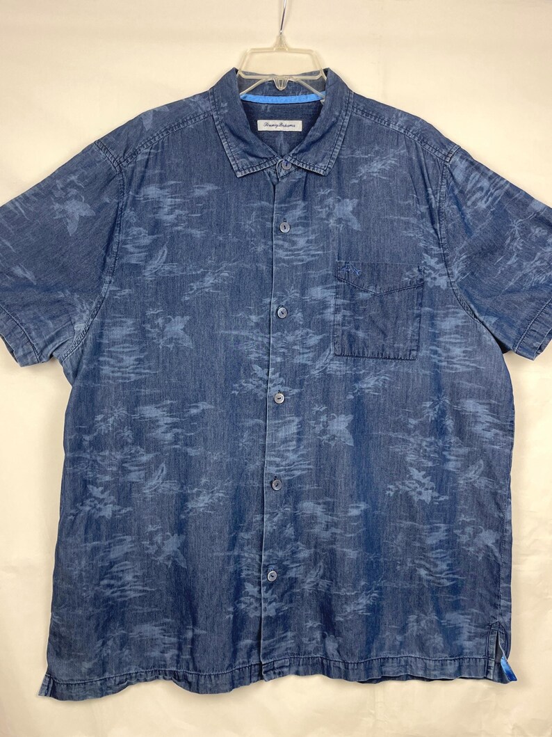 Może przedstawiać: Koszula dżinsowa z kr&oacute;tkim rękawem w średnim niebieskim odcieniu, z subtelnym kwiatowym wzorem w jaśniejszym odcieniu niebieskiego. Posiada jedną kieszeń na piersi i klasyczny kołnierzyk. Koszula jest zapięta i wisi na białym wieszaku.