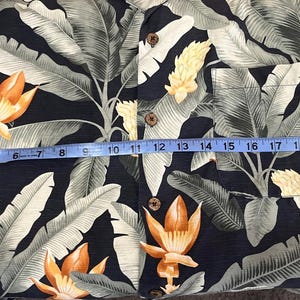 Puede incluir: Una camisa con botones con un estampado floral tropical. La camisa presenta un fondo azul marino oscuro con hojas grandes de color verde claro y flores naranjas y amarillas. La longitud de la camisa es de aproximadamente 61 cm.