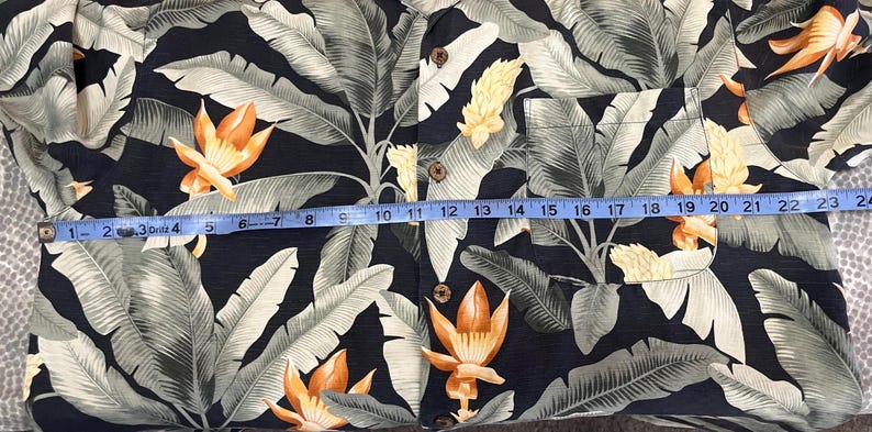 Puede incluir: Una camisa con botones con un estampado floral tropical. La camisa presenta un fondo azul marino oscuro con hojas grandes de color verde claro y flores naranjas y amarillas. La longitud de la camisa es de aproximadamente 61 cm.