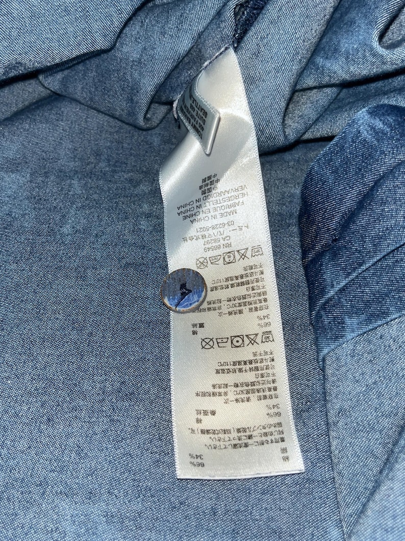 Może przedstawiać: Zbliżenie niebieskiego materiału dżinsowego z dołączoną białą metką. Etykieta zawiera tekst w wielu językach, w tym chińskim, japońskim i angielskim, wraz z symbolami pielęgnacji. Do materiału przymocowany jest guzik.