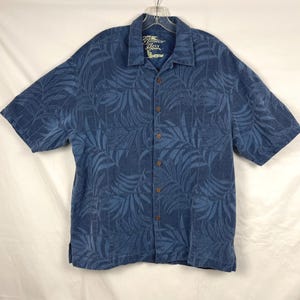 Puede incluir: Camisa azul de manga corta con un estampado de hojas tropicales. La camisa tiene cuello y botones de madera. El nombre de la marca "Jamaica Jax" es visible en la etiqueta.