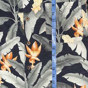 Puede incluir: Una camisa con botones con un fondo azul oscuro y un estampado floral tropical. La camisa presenta hojas grises grandes y flores naranjas y amarillas. La camisa mide aproximadamente 74 cm de largo.