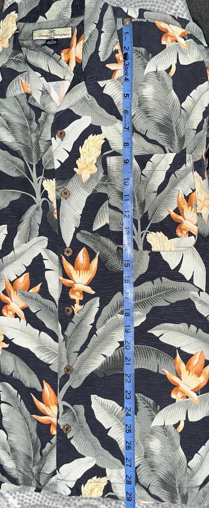 Puede incluir: Una camisa con botones con un fondo azul oscuro y un estampado floral tropical. La camisa presenta hojas grises grandes y flores naranjas y amarillas. La camisa mide aproximadamente 74 cm de largo.