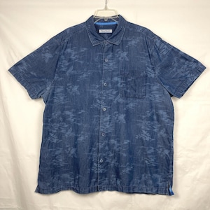 Może przedstawiać: Niebieska koszula z kr&oacute;tkim rękawem zapinana na guziki z subtelnym, całopowierzchniowym wzorem. Koszula ma kieszeń na piersi i klasyczny kołnierzyk. Nazwa marki "Tommy Bahama" jest widoczna na kołnierzyku. Koszula wisi na białym wieszaku.