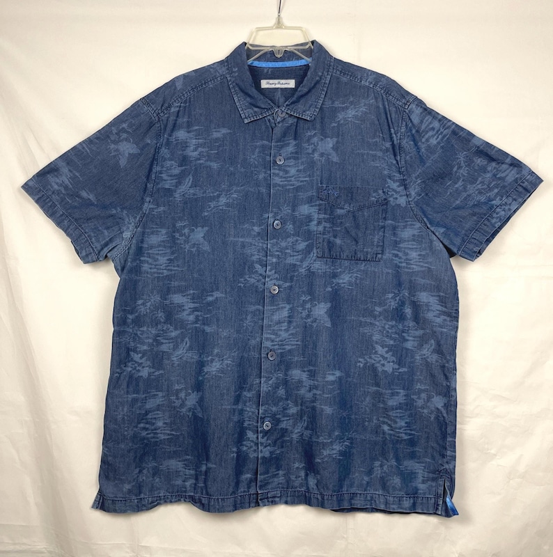 Może przedstawiać: Niebieska koszula z kr&oacute;tkim rękawem zapinana na guziki z subtelnym, całopowierzchniowym wzorem. Koszula ma kieszeń na piersi i klasyczny kołnierzyk. Nazwa marki "Tommy Bahama" jest widoczna na kołnierzyku. Koszula wisi na białym wieszaku.