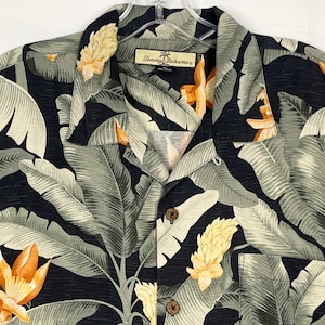 Puede incluir: Una camisa negra con un estampado floral tropical. La camisa presenta hojas grandes de color verde claro y flores naranjas sobre un fondo oscuro. Se ve la marca "Tommy Bahama".