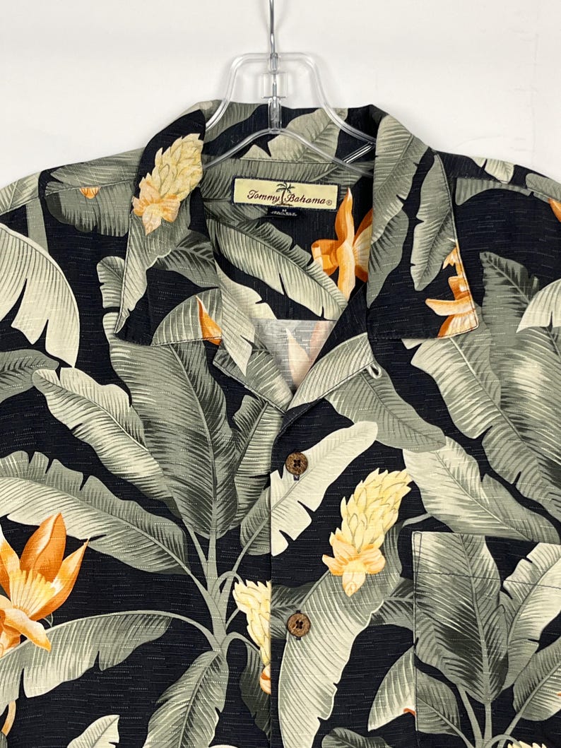 Puede incluir: Una camisa negra con un estampado floral tropical. La camisa presenta hojas grandes de color verde claro y flores naranjas sobre un fondo oscuro. Se ve la marca "Tommy Bahama".