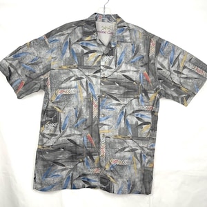 May include: A gray, short-sleeved button-down shirt with a colorful abstract print of leaves and geometric patterns. The shirt has a collar and a button closure. The brand name "Bamboo Cay" is visible on the collar.