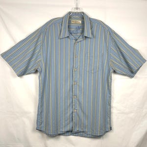 Puede incluir: Camisa abotonada de manga corta a rayas azules y amarillas. La camisa tiene un bolsillo en el pecho y un cuello. El nombre de la marca est&aacute; bordado en el cuello. La camisa est&aacute; colgada de una percha.