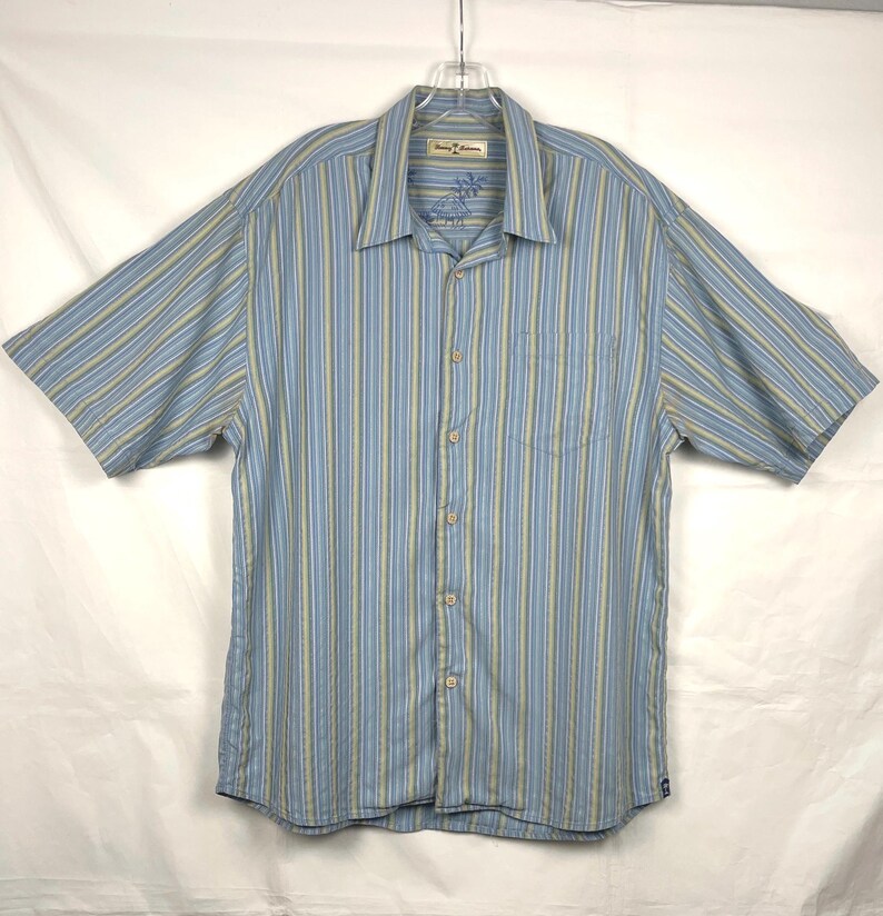 Puede incluir: Camisa abotonada de manga corta a rayas azules y amarillas. La camisa tiene un bolsillo en el pecho y un cuello. El nombre de la marca est&aacute; bordado en el cuello. La camisa est&aacute; colgada de una percha.