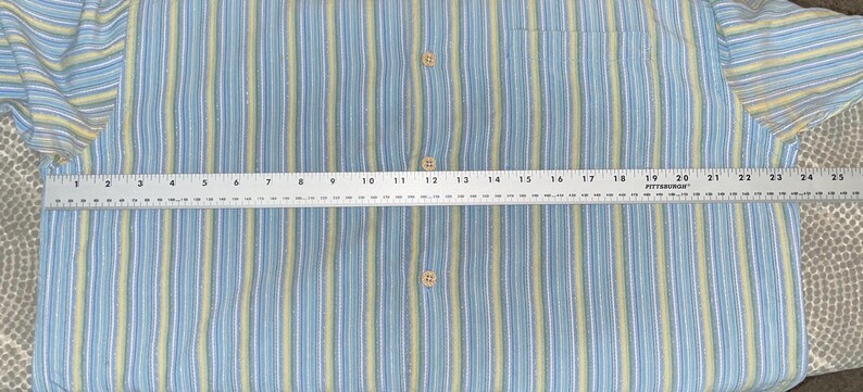 Puede incluir: Camisa abotonada a rayas azules y amarillas con bolsillo en el pecho. La camisa se mide con una regla, mostrando un ancho de aproximadamente 63,5 cm. La camisa tiene un estilo cl&aacute;sico e informal.