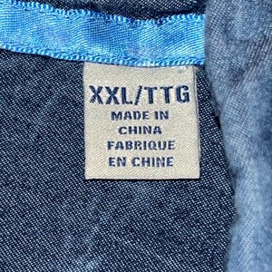 Może przedstawiać: Zbliżenie niebieskiej koszuli dżinsowej z jasnoniebieskim kołnierzem. Biała metka z czarnym tekstem głosi "XXL/TTG MADE IN CHINA FABRIQUE EN CHINE". Tkanina koszuli ma teksturowany wygląd.