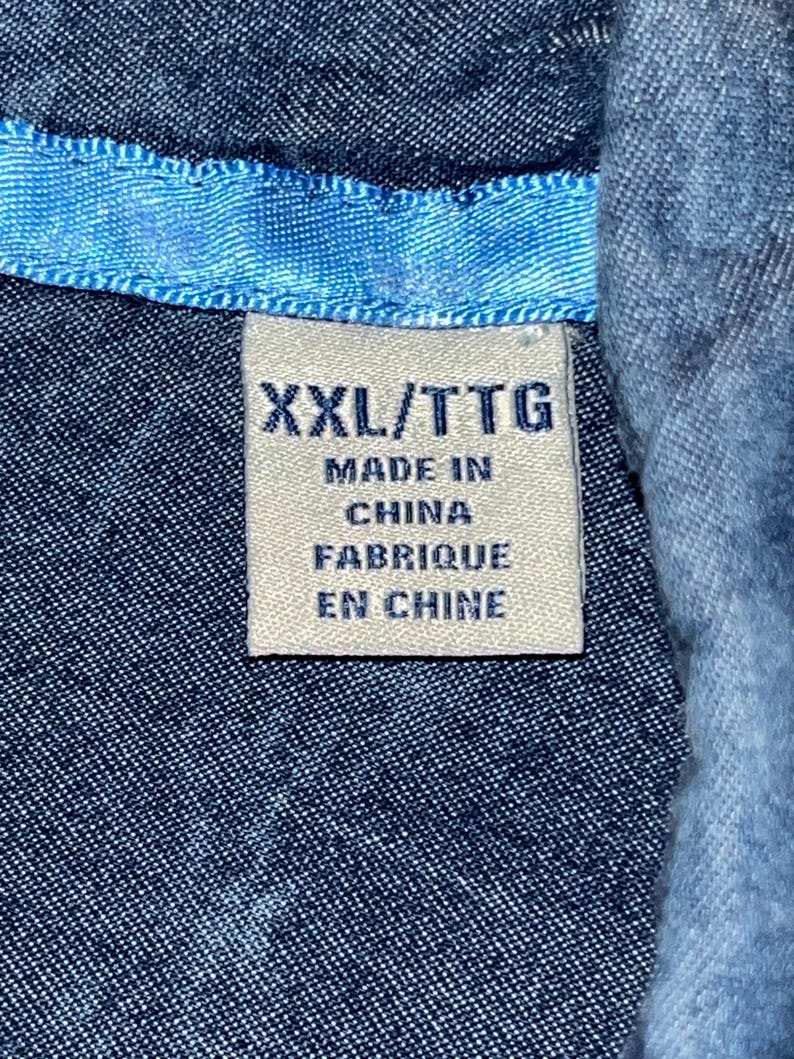Może przedstawiać: Zbliżenie niebieskiej koszuli dżinsowej z jasnoniebieskim kołnierzem. Biała metka z czarnym tekstem głosi "XXL/TTG MADE IN CHINA FABRIQUE EN CHINE". Tkanina koszuli ma teksturowany wygląd.