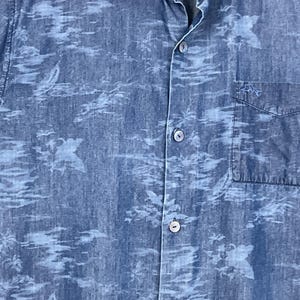 Może przedstawiać: Niebieska koszula z jasnoniebieskim wzorem. Koszula ma kieszeń na piersi i kołnierzyk. Materiał wydaje się być dżinsem lub podobnym. Koszula jest rozpięta u g&oacute;ry.