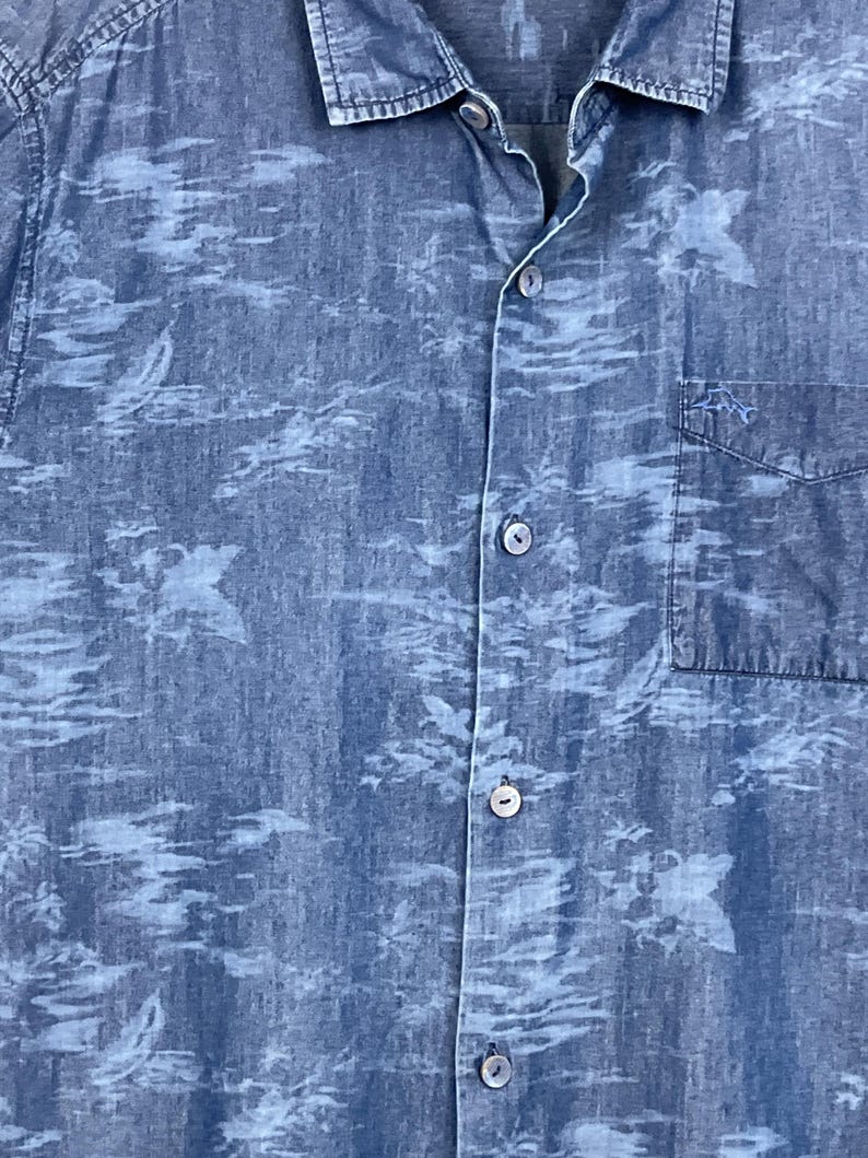 Może przedstawiać: Niebieska koszula z jasnoniebieskim wzorem. Koszula ma kieszeń na piersi i kołnierzyk. Materiał wydaje się być dżinsem lub podobnym. Koszula jest rozpięta u g&oacute;ry.