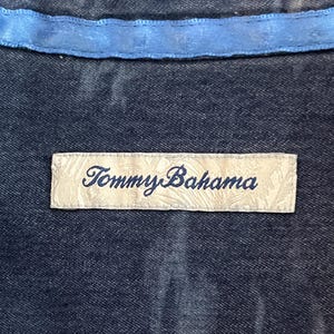 Może przedstawiać: Zbliżenie na ciemnoniebieską tkaninę z jasnoniebieskim wykończeniem. Prostokątna metka z napisem "Tommy Bahama" w kursywie jest wszyta w tkaninę. Etykieta jest w kolorze kremowym.