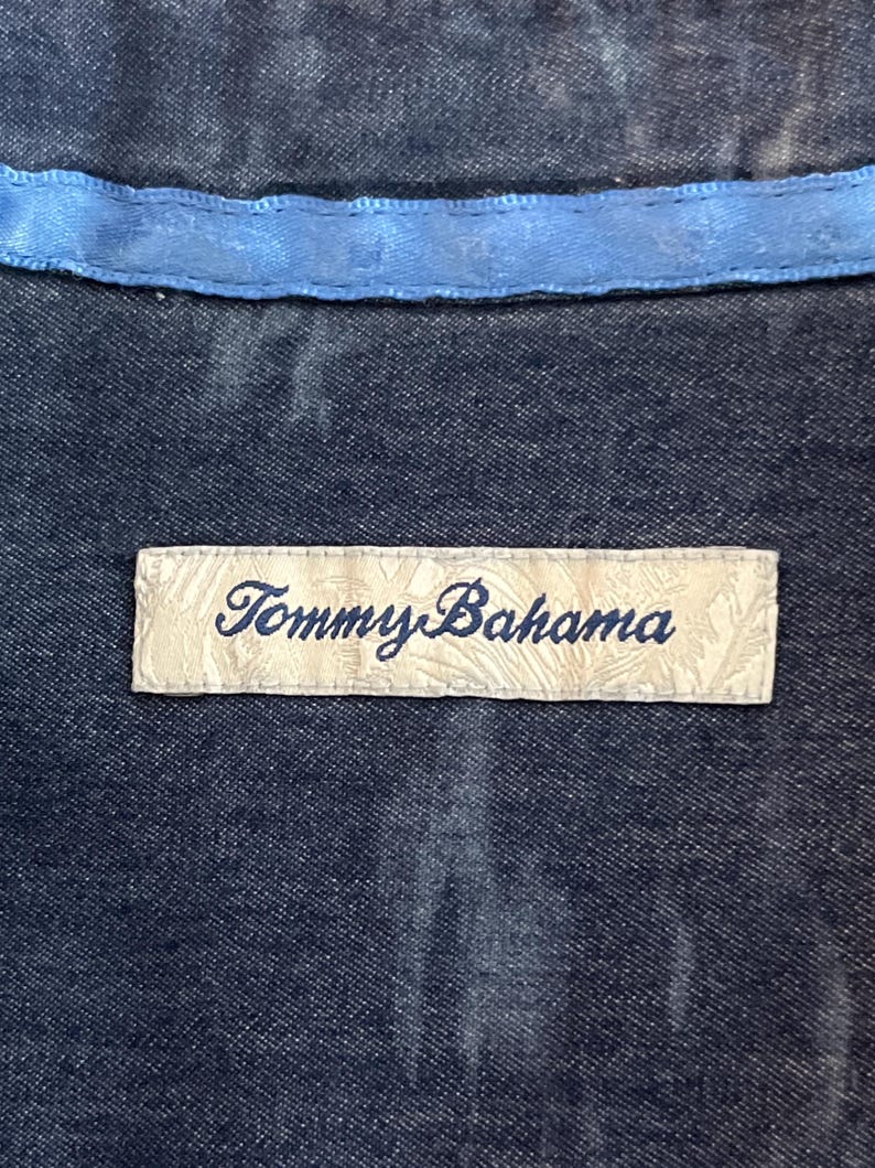 Może przedstawiać: Zbliżenie na ciemnoniebieską tkaninę z jasnoniebieskim wykończeniem. Prostokątna metka z napisem "Tommy Bahama" w kursywie jest wszyta w tkaninę. Etykieta jest w kolorze kremowym.