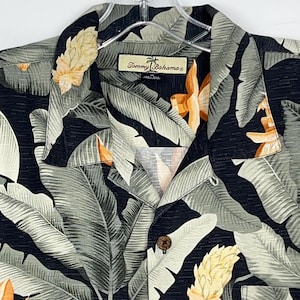 Puede incluir: Una camisa negra con un estampado de hojas y flores tropicales. La camisa presenta grandes hojas grises y blancas con flores amarillas y naranjas. La etiqueta de la marca "Tommy Bahama" es visible en el cuello.