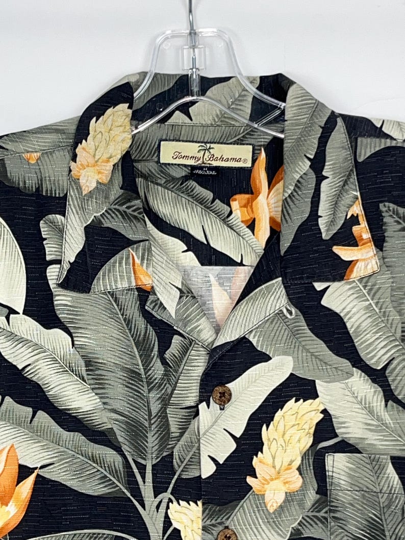 Puede incluir: Una camisa negra con un estampado de hojas y flores tropicales. La camisa presenta grandes hojas grises y blancas con flores amarillas y naranjas. La etiqueta de la marca "Tommy Bahama" es visible en el cuello.