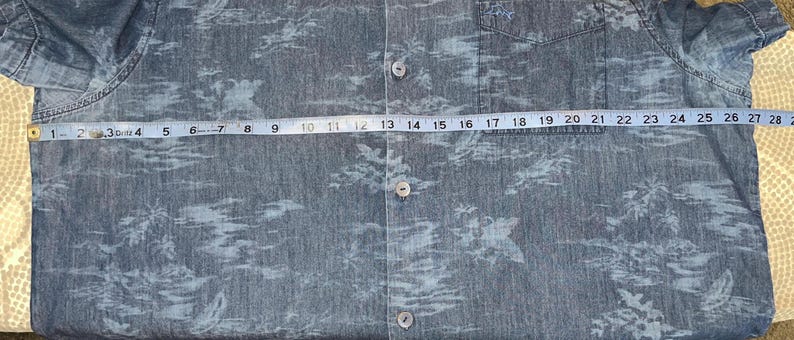 Może przedstawiać: Niebieska koszula zapinana na guziki z tropikalnym nadrukiem. Koszula jest mierzona taśmą mierniczą, pokazując szerokość około 71 cm. Koszula ma kieszeń na lewej piersi i kr&oacute;tkie rękawy.