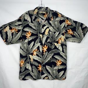 Puede incluir: Una camisa de manga corta con botones y un estampado floral tropical. La camisa presenta un fondo azul marino oscuro con hojas verdes grandes y flores naranjas y amarillas. Tiene un bolsillo en el pecho y botones de madera.