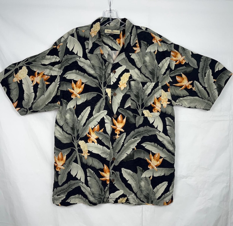 Puede incluir: Una camisa de manga corta con botones y un estampado floral tropical. La camisa presenta un fondo azul marino oscuro con hojas verdes grandes y flores naranjas y amarillas. Tiene un bolsillo en el pecho y botones de madera.