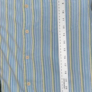 Puede incluir: Camisa abotonada con rayas verticales en azul claro, amarillo y blanco. La camisa tiene un bolsillo en el pecho y mide aproximadamente 76 cm de largo. La marca "Tommy Bahama" es visible en el cuello.